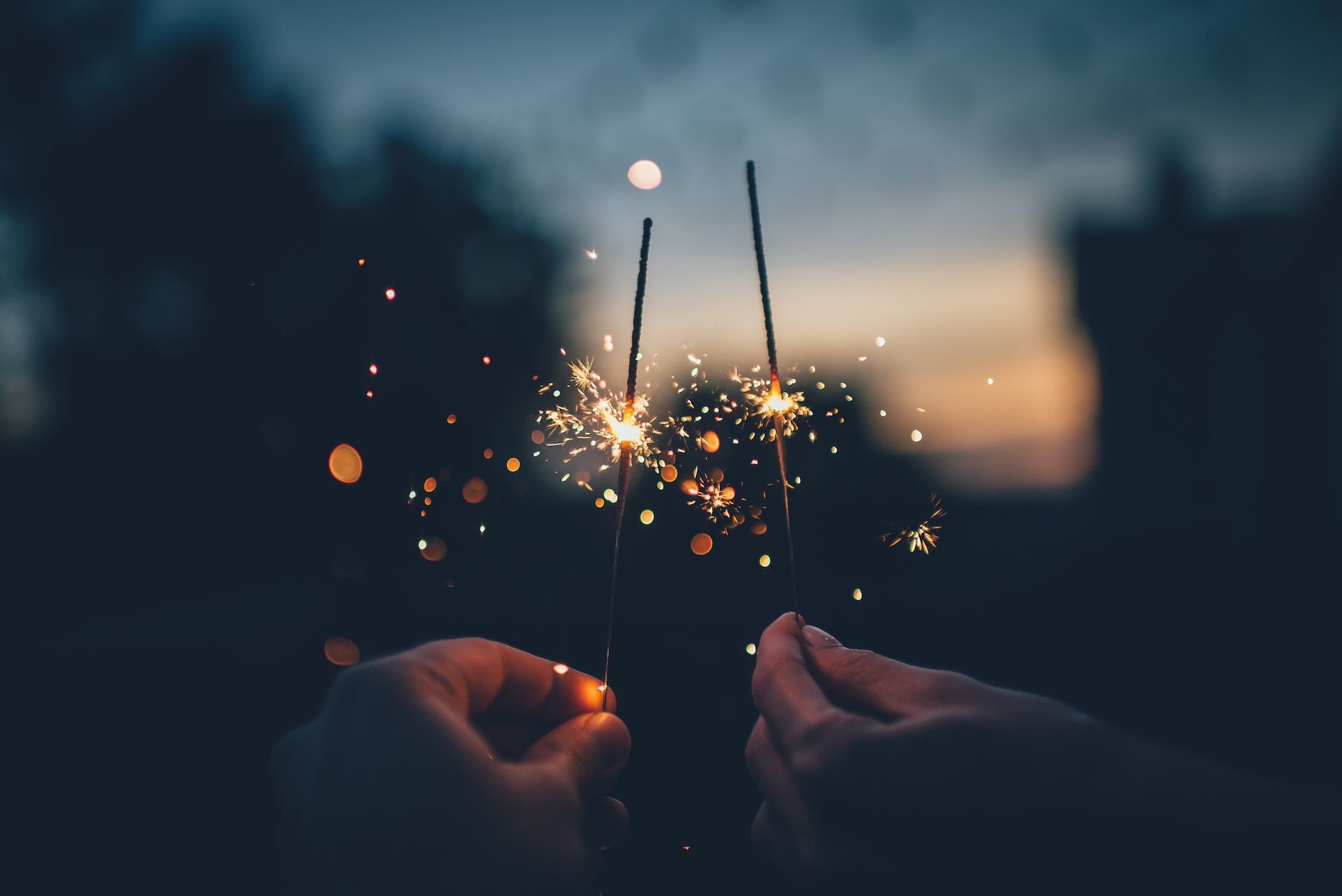 two hands holding sparklers against silhouettes of buildings at dusk