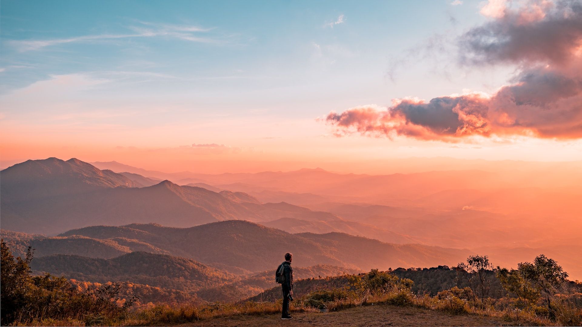 man standing atop a hill overlooking a range of hills against a dawn-orange sky