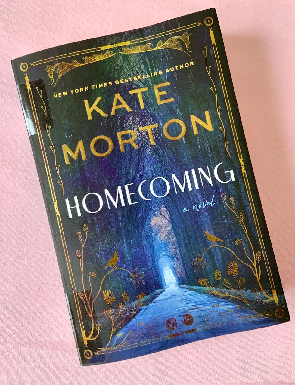 Paperback copy of Homecoming by Kate Morton, featuring a trail through the woods with a blue light at the end