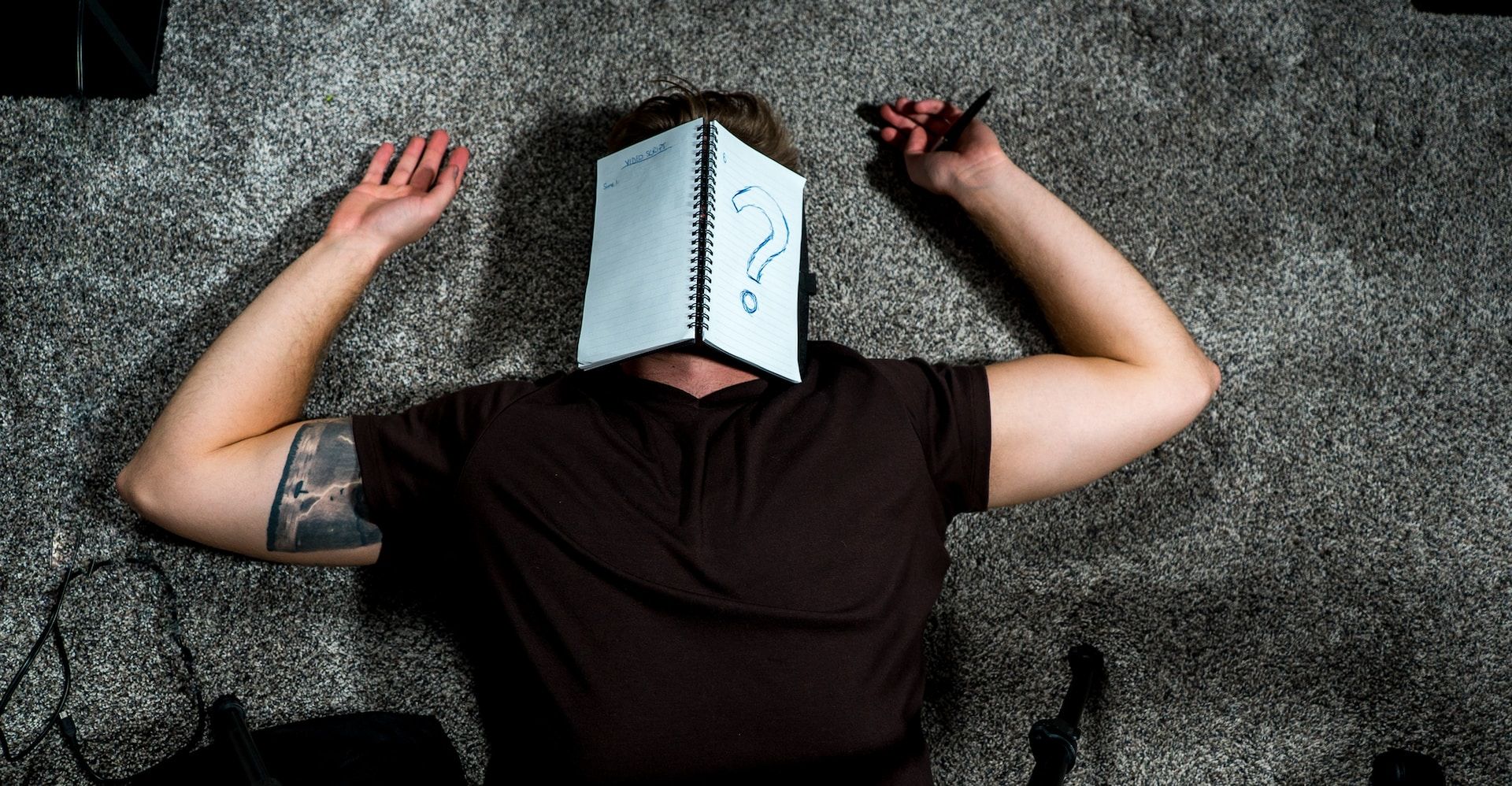 man lying on the ground with an open spiral-bound notebook covering his face and a question mark on the page