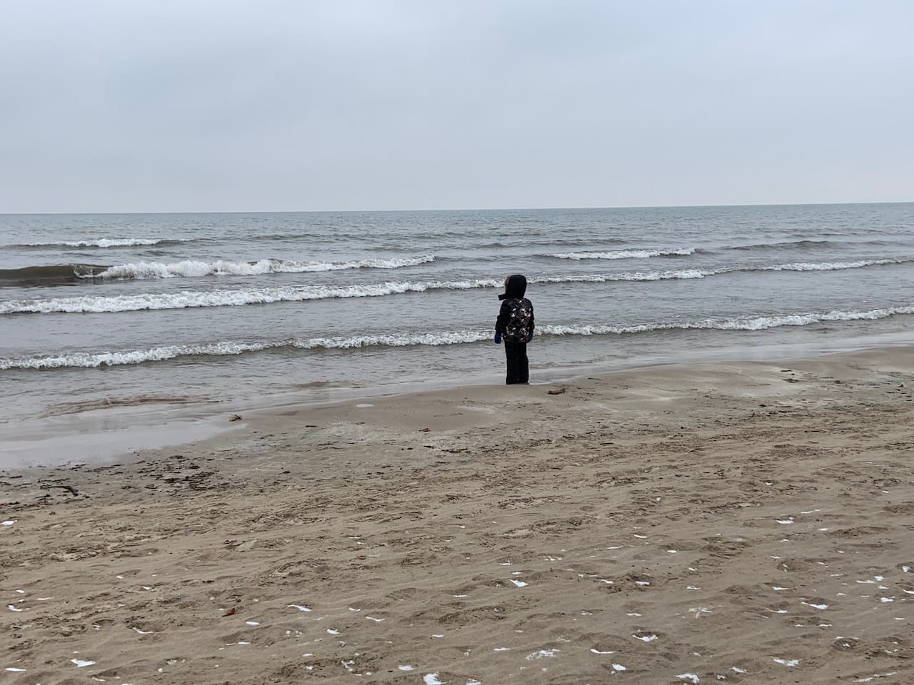 a child in winter gear standing on the beach and looking out at the waves