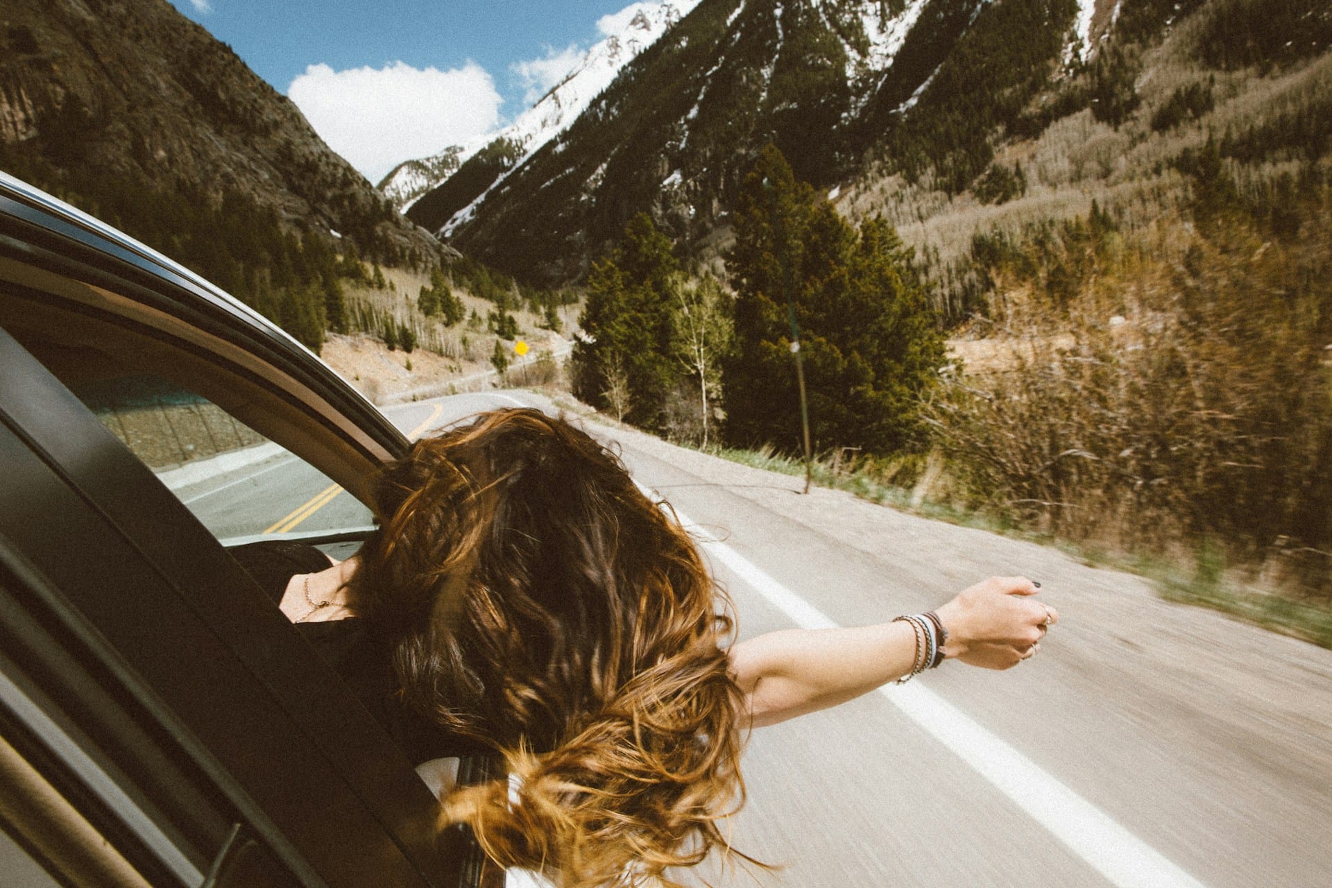woman leaning out of a car window and enjoying the wind on her face on a road flanked by mountains