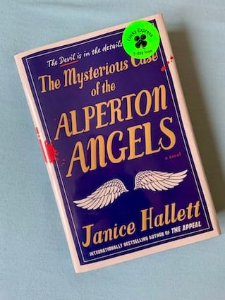 hardback copy of The Mysterious Case of The Alperton Angels by Janice Hallett featuring a pair of disembodied angel wings against a dark blue background