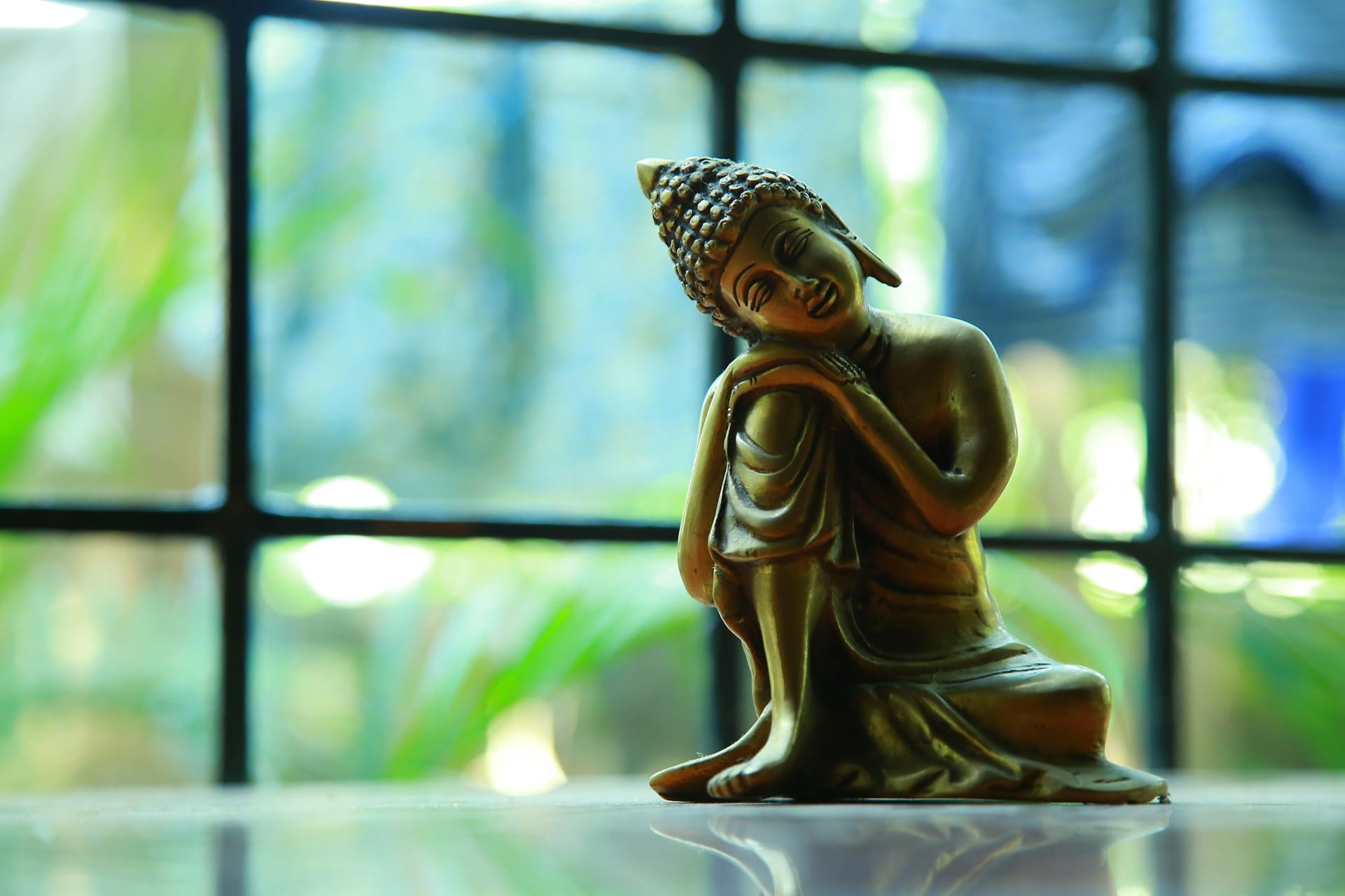 statue of a Buddha resting his head on his knee against a blurred backdrop of a window overlooking a garden