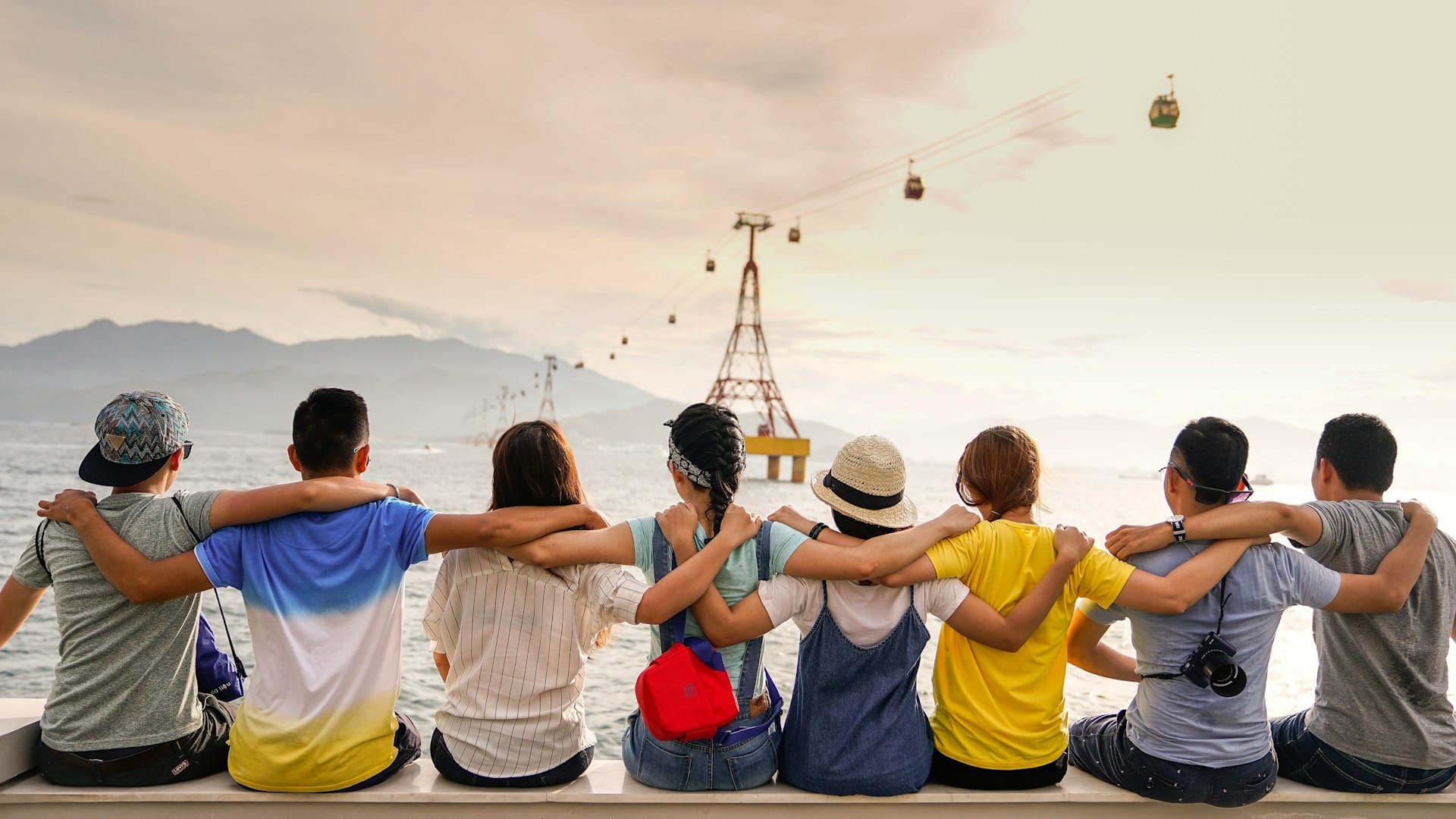 a group of friends sitting on a ledge and looking out at a water body with silhouettes of hills in the distance and a cable car ride