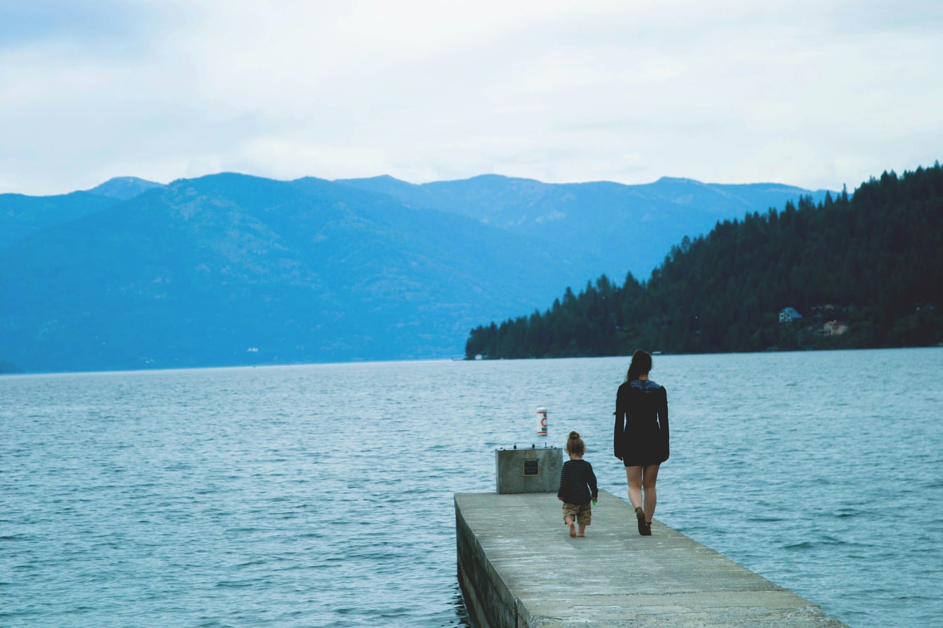 an adult woman and a small child on a boardwalk pier on a body of water with hills in the distance