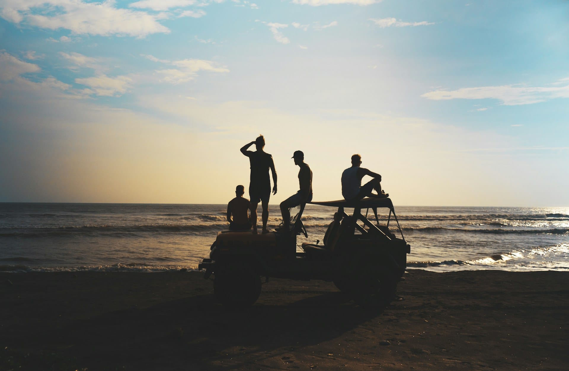 silhouette of a group of young men atop an SUV parked by the seashore looking out towards the ocean