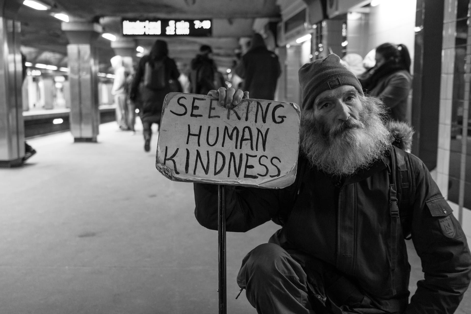bearded man holding a board that says 'Seeking Human Kindness' on a railway platform