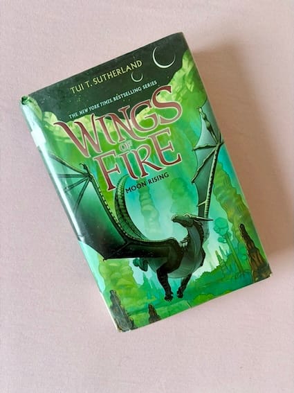 hardback copy of Moon Rising by Tui T. Sutherland featuring a dark dragon against the backdrop of a green forest