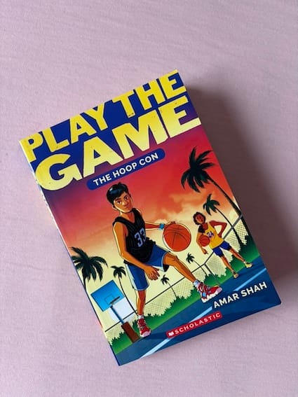 paperback copy of The Hoop Con by Amar Shah featuring an Indian boy and girl with basketballs on a court surrounded by palm trees