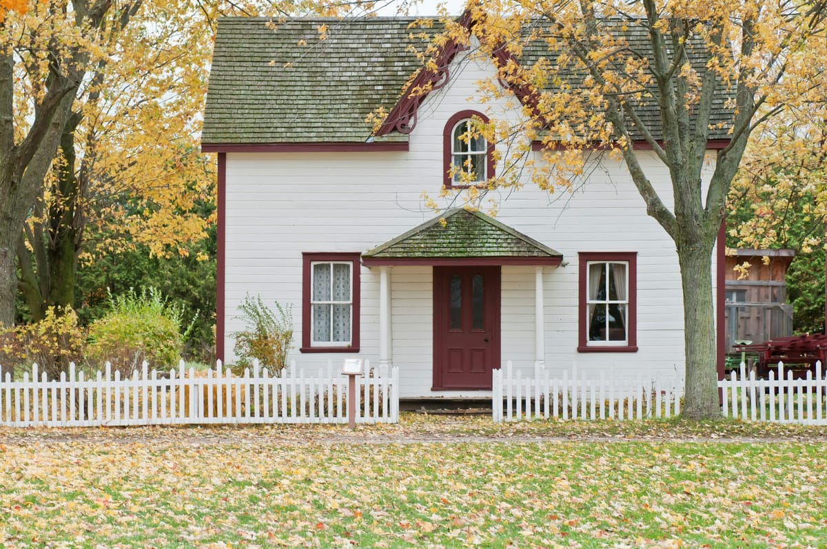 a white house with red trims under maple trees with leaves turning yellow in autumn