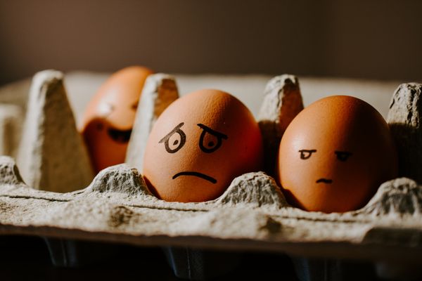 three eggs in a carton with faces painted on them, one annoyed, another sad, and the third laughing