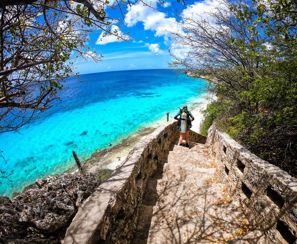 a backpacker standing on steps leading down to a sandy beach by a blue-green ocean under a bright blue sky