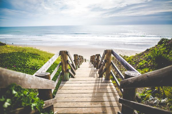 wooden walkway with steps leading down to a sandy beach with pale blue waters under a bright sky
