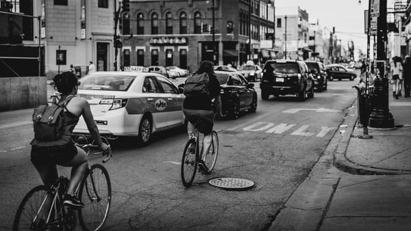 black and white image of cyclists and cars on a busy city street flanked by buildings and sidewalks