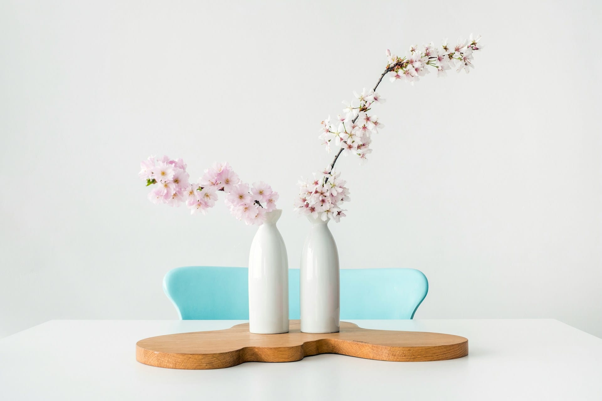 flowers in two vases atop a wooden coaster on a white table with a turquoise blue chair behind