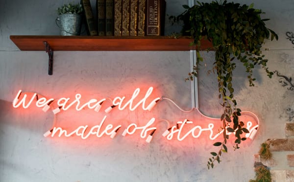 a neon wall sign reading 'We are all made of stories' under a shelf with books and a potted plant