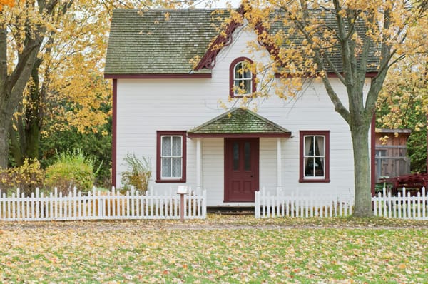 a white house with red trims under maple trees with leaves turning yellow in autumn