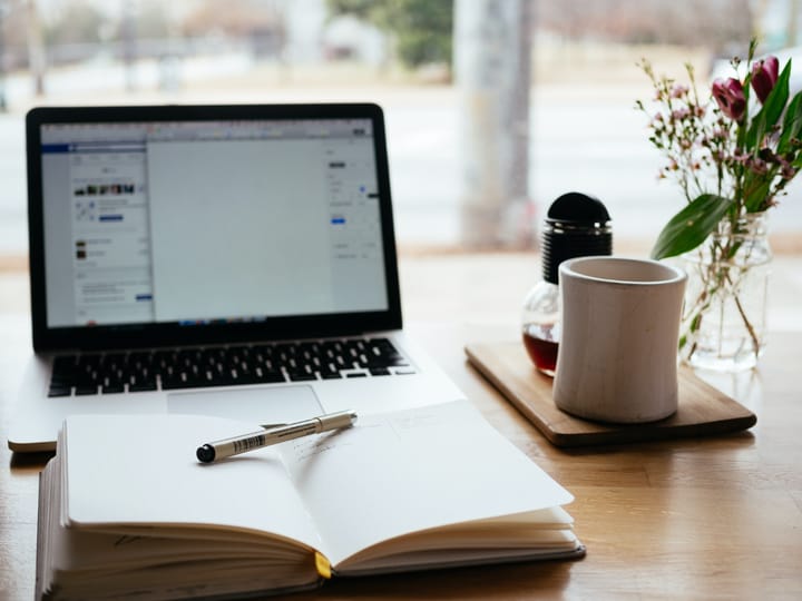 a laptop, an open notebook, a coffee cup on a coaster, and a sprig of flowers in a glass jar on a table