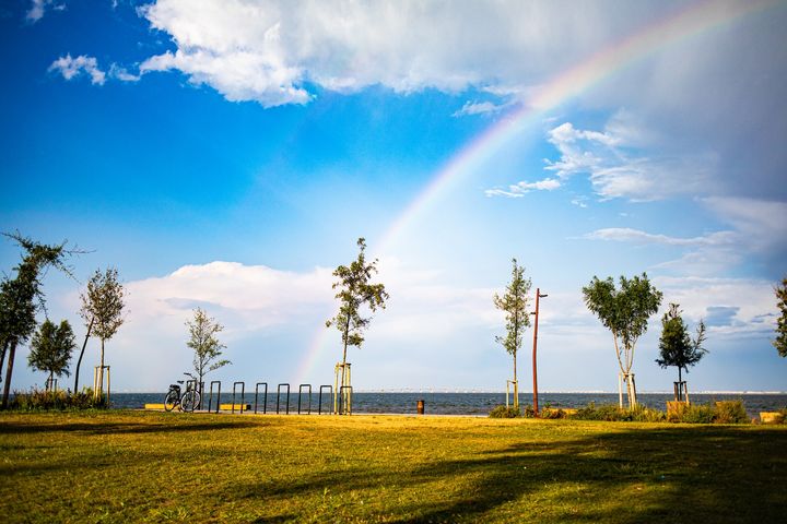 green grass field and scattered trees beside a river, all under a blue sky with white clouds and a rainbow