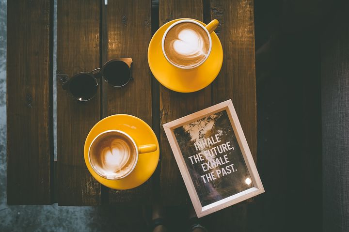 two café lattes in yellow cups and saucers with a pair of sunglasses and a picture frame on a wooden table top