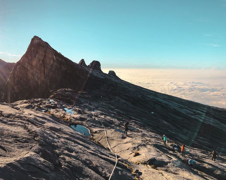 climbers making their way up a mountain slop towards the peak with a sea of clouds and the blue sky in the background