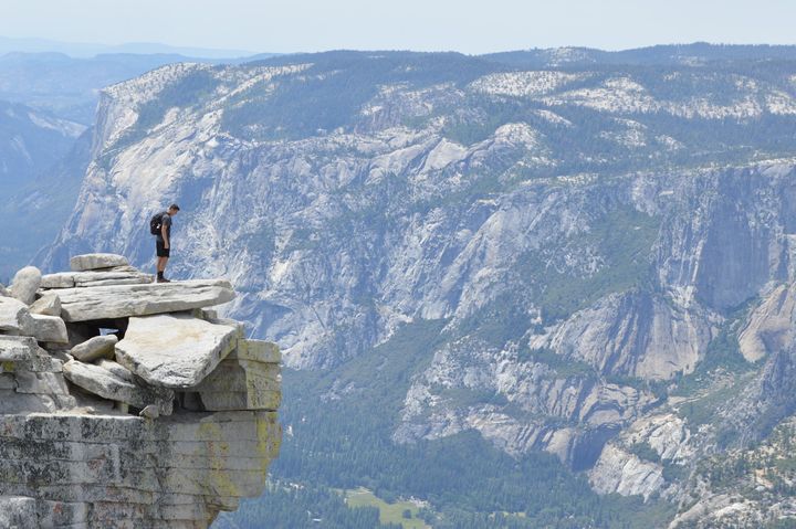 man on a ledge atop a cliff against a backdrop of a steep mountain range