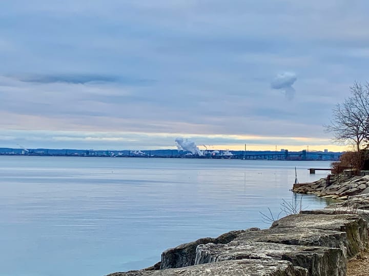 clouds of smoke rising from factories on the far border of a blue lake under a blue sky