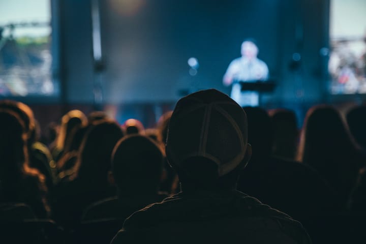 blurred image of a room full of people facing a speaker on a stage