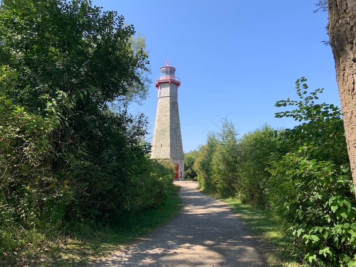 a path leading to a defunct lighthouse, flanked by bushes on both sides, under a bright blue sky