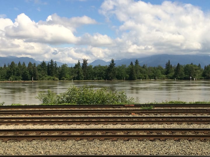 train tracks running along the banks of a lake with evergreen trees on the other bank and mountains in the far distance