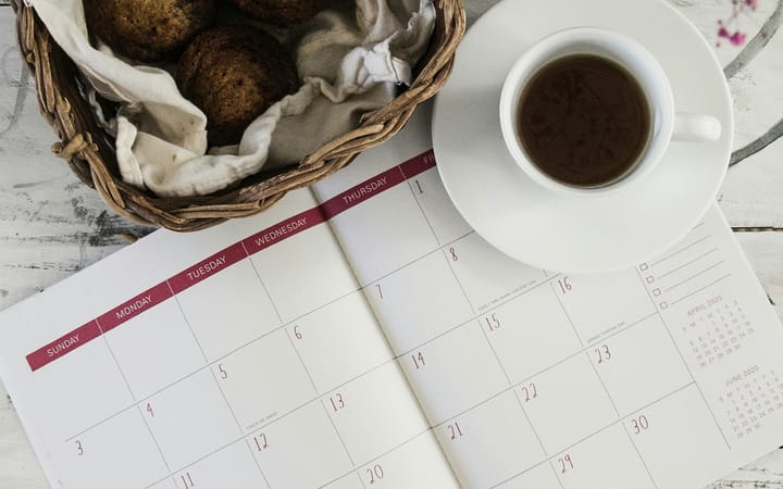 a planner opened to a monthly schedule page, a cup of black coffee, and a basket of bread on a white table