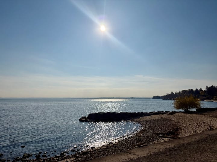 sun shining in a pale blue sky above the waters of Lake Ontario flanked by a curved strip of beach on the right side